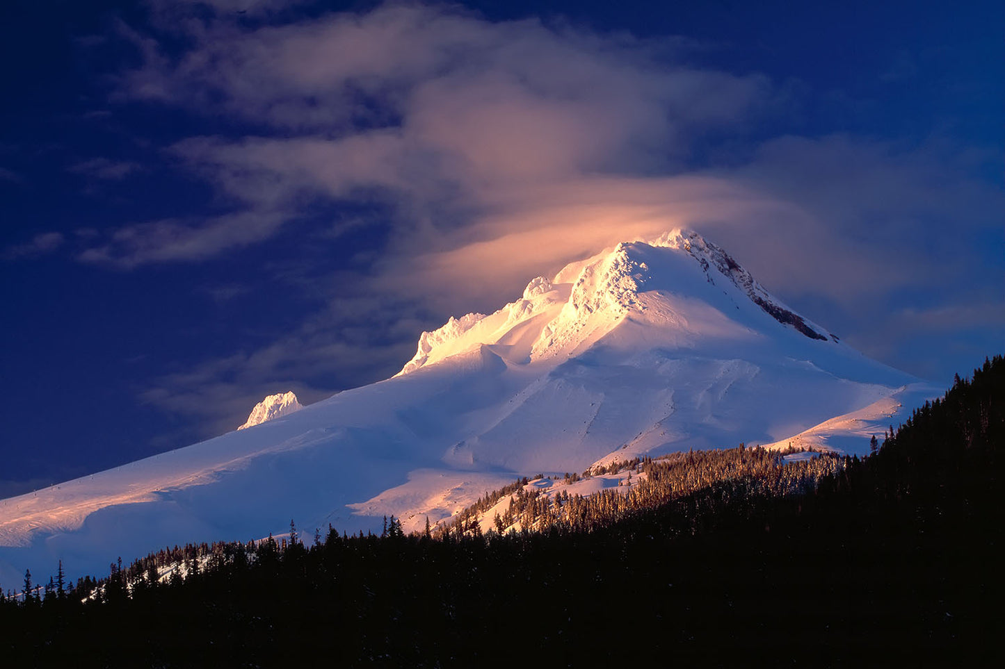 Sun illuminates the summit of snow covered Mt. Hood. Clouds rise off the peak and surrounding forest in Oregon.