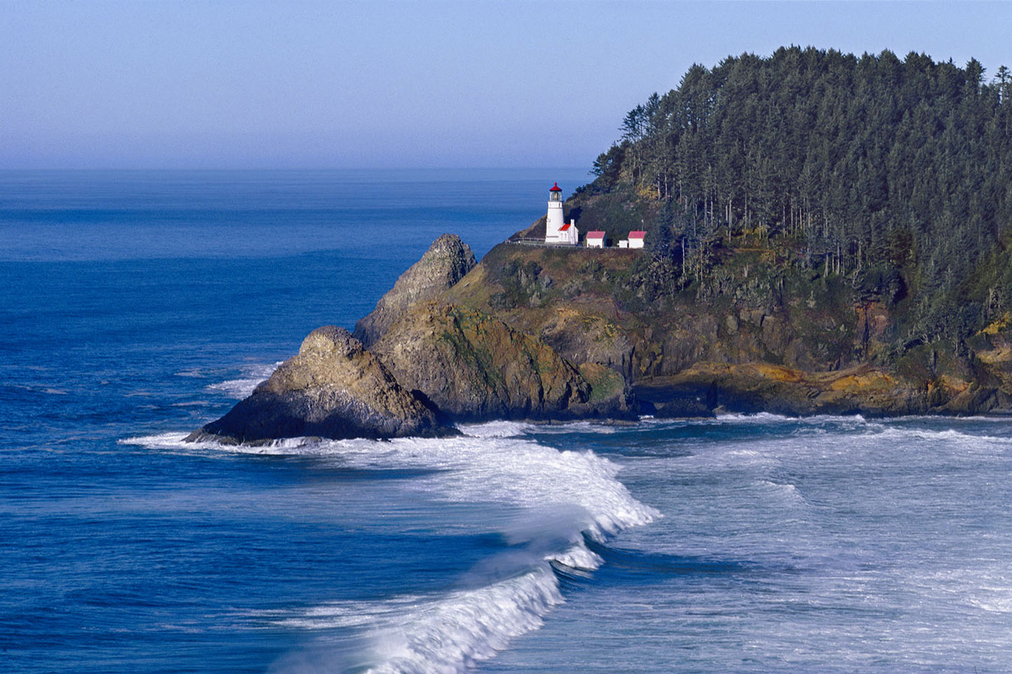 Heceta Head Lighthouse stands high on a forested cliff. Waves of the Pacific Ocean roll past on a sunny day in Florence, Oregon.