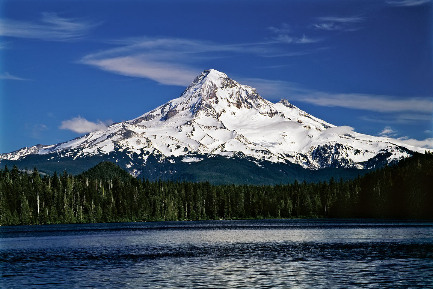 Snow covered Mt. Hood stands tall against a blue sky. Green forest surrounds the rippled waters of Lost Lake, Oregon below.