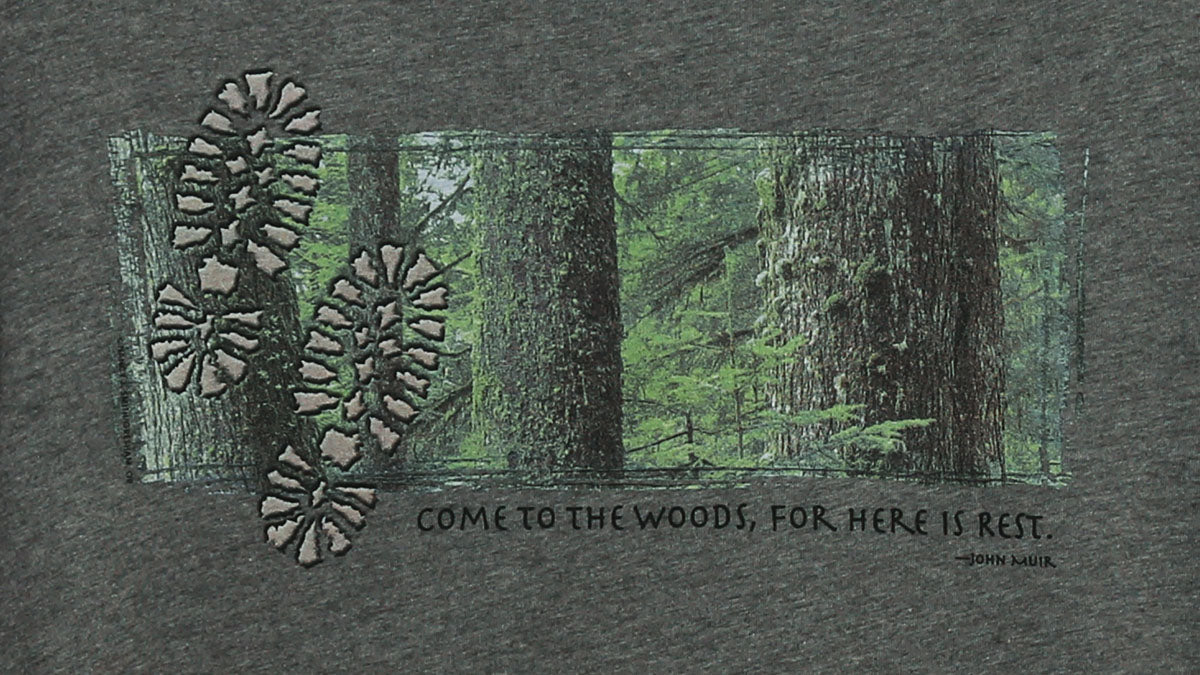 Detail view of shirt print on dark heather grey. Features tree trunks against forest background with two footprints on left side. Caption states "Come to the woods, for here is rest."