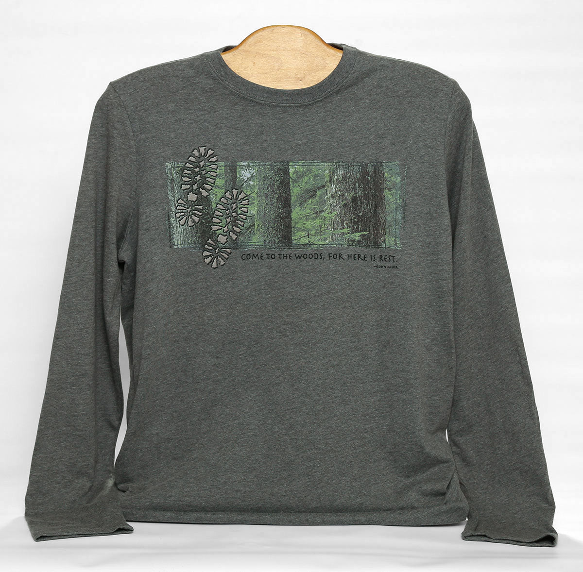 Front view of Come to the woods long sleeve t-shirt in color dark heather grey. Print features tree trunks in a forest with two footprints and caption "Come to the woods, for here is rest."
