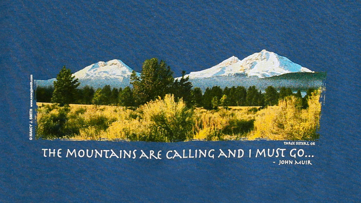 Detail view of shirt screen print on indigo blue, featuring Oregon's Three sisters mountains and caption "The Mountains are Calling and I Must Go...".