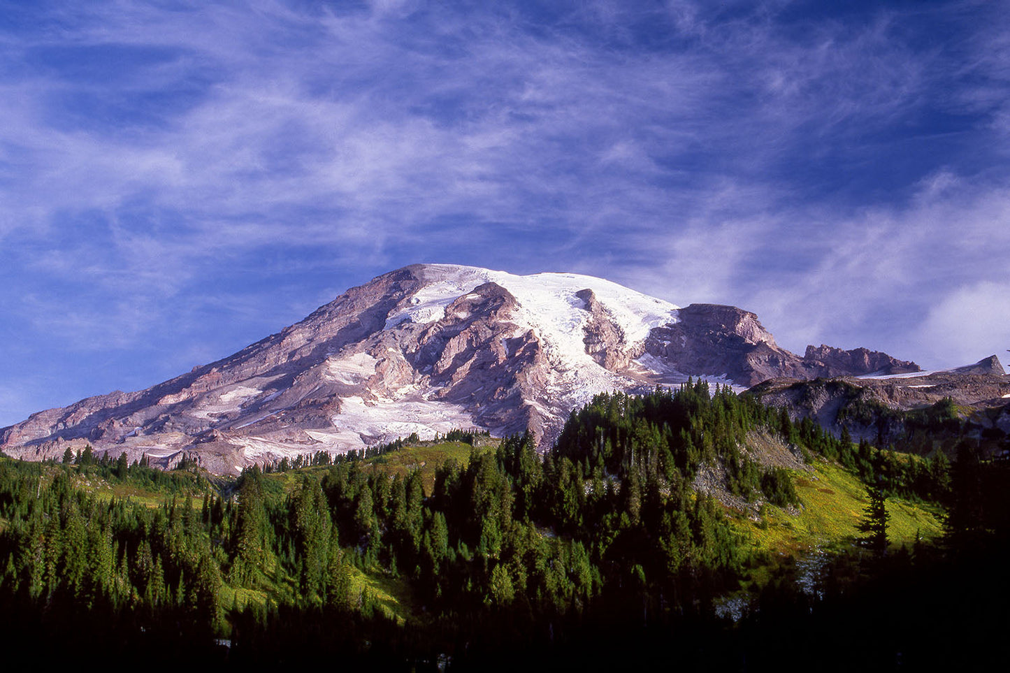 Snow dusted Mt. Rainier stands tall into wispy clouds and a blue sky. Green forested Washington foothills surround the base of the mountain.