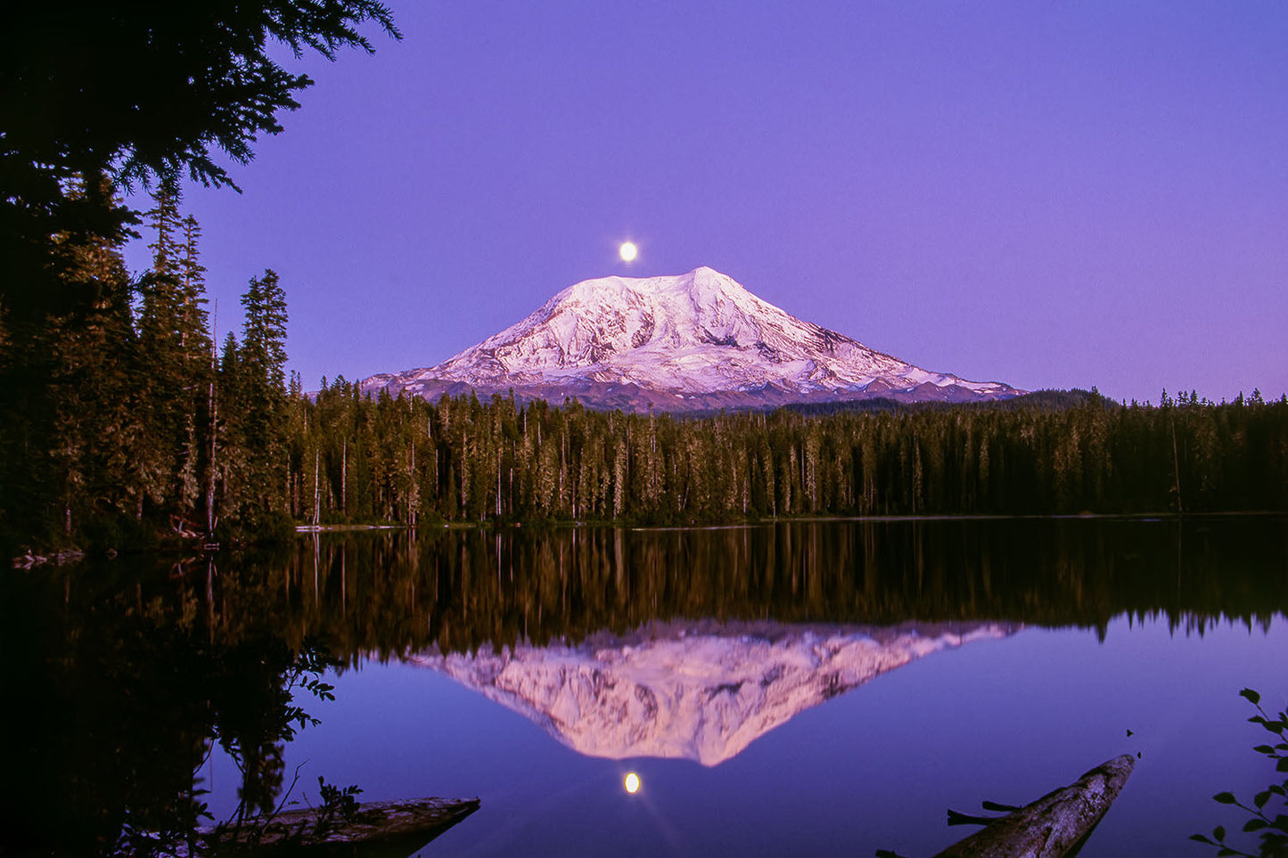 A full moon rises into a clear purple sky over Mt. Adams in Gifford Pinchot National Forest, Washington State. Tahklak lake below, surrounded by forest, reflects the mountain.