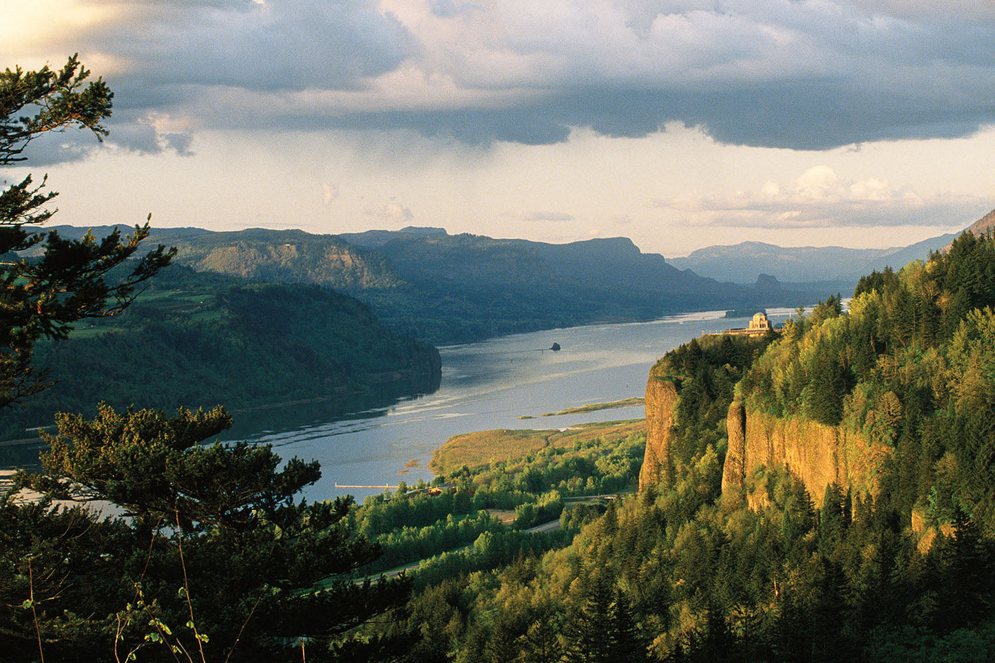 Sun illuminates Crown Point, with Vista House sitting atop. Green forest lays in the surrounding area, as the Columbia River runs calmly through the Gorge in Oregon.