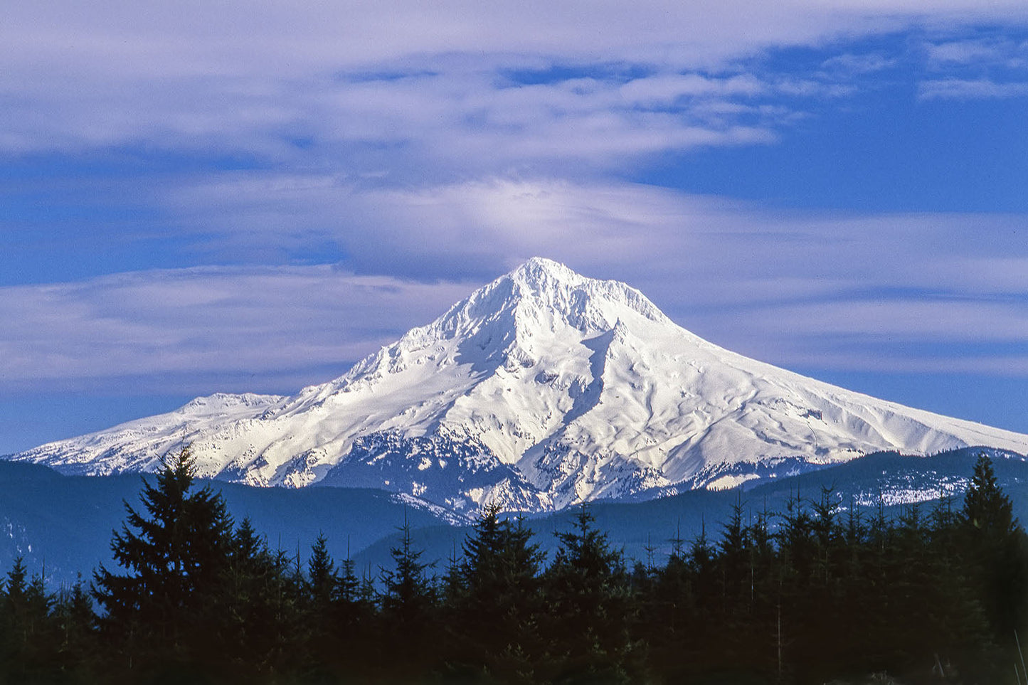 Snow covered Mt. Hood stands tall over green forested foothills, protruding into a cloudy blue Oregon sky.