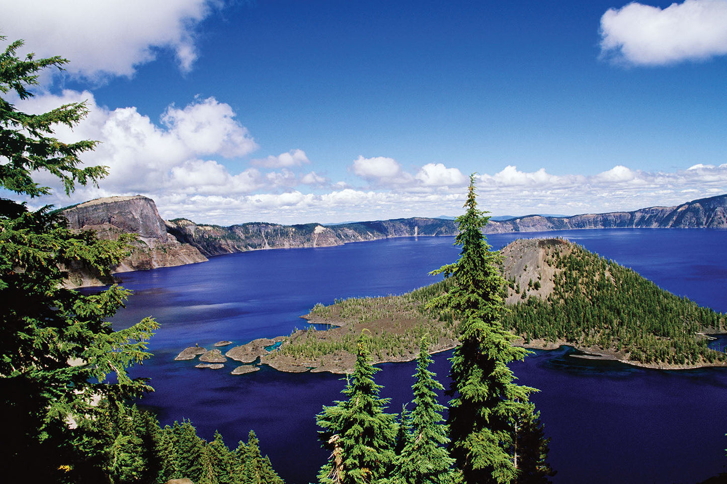 Wizard Island sits upon the deep blue waters of Crater Lake, Oregon. Blue skies engulf smooth rocky cliffs in the distance.