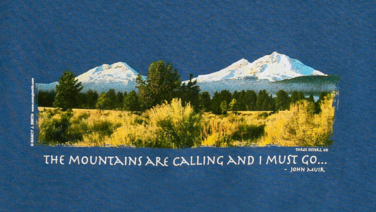 Detail view of shirt screen print on indigo blue, featuring Oregon's Three sisters mountains and caption "The Mountains are Calling and I Must Go...".